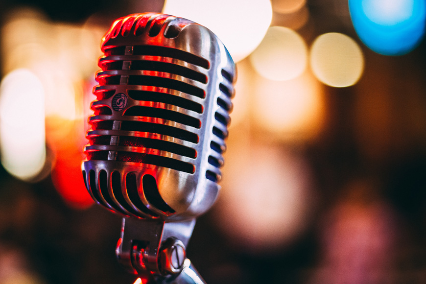 A silver microphone in focus against a blurry background. There are out of focus yellow, red, and blue lights in indistinguishable shapes in the background. 